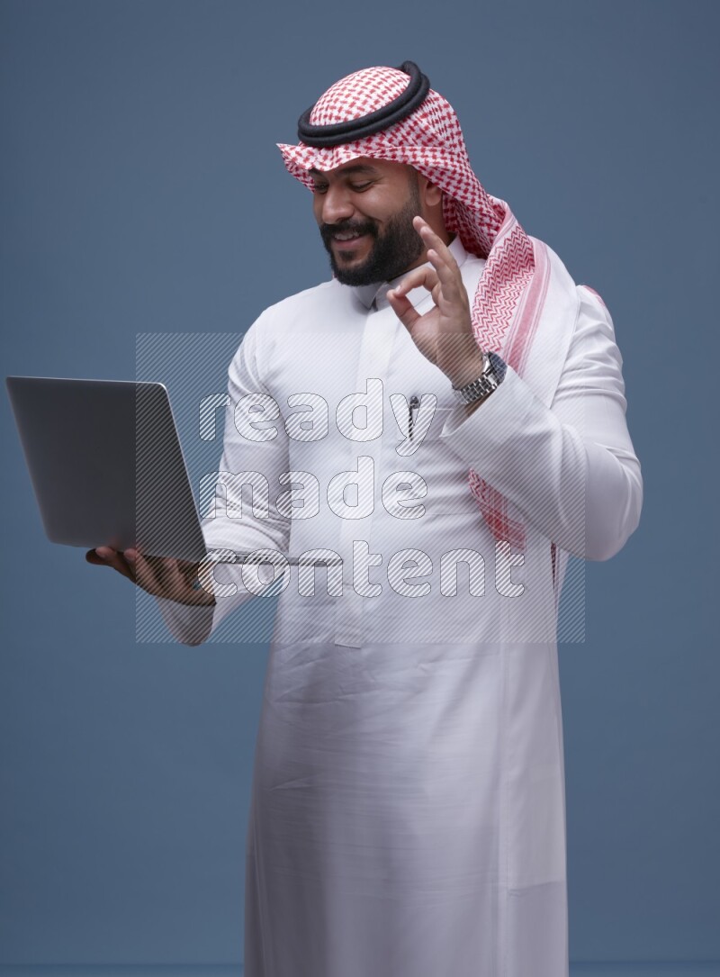 A man standing  with a laptop on Blue Background wearing Saudi Thob and Shomag
