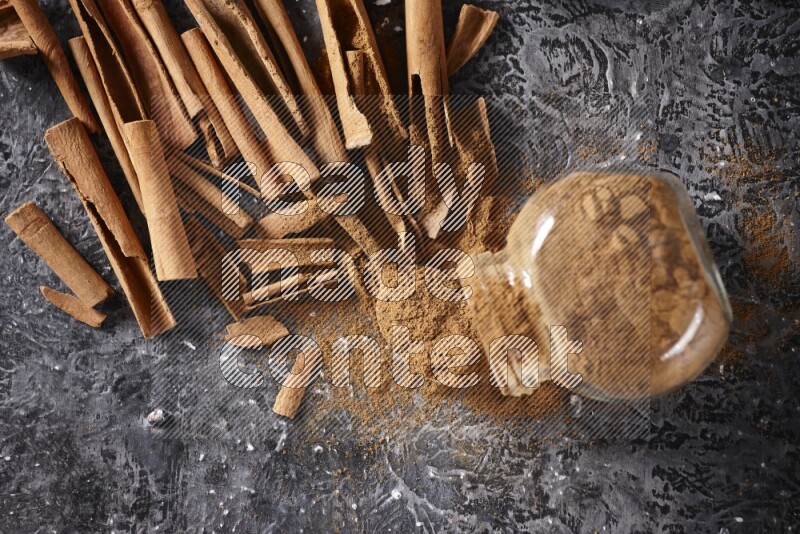 Herbal glass jar full cinnamon powder flipped and a metal spoon full of powder surrounded by cinnamon sticks on textured black background in different angles