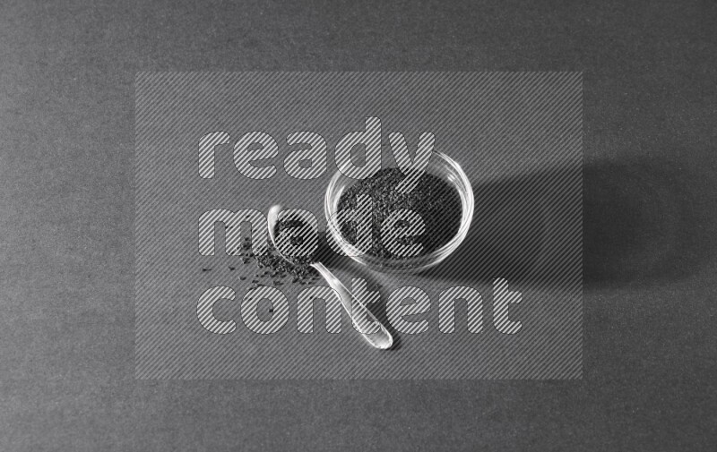 A glass bowl full of black seeds with a metal spoon full of the seeds on a black flooring