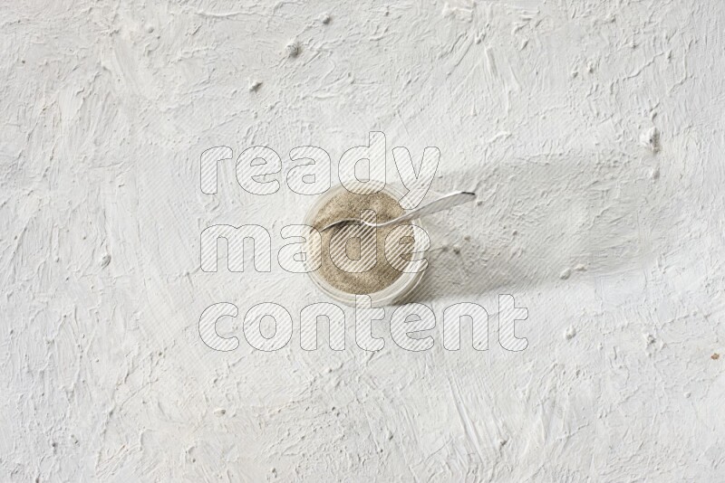 A glass jar and a metal spoon full of white pepper powder on textured white flooring