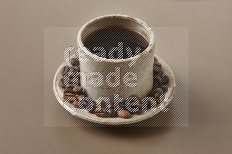 A beige pottery cup of coffee surrounded by roasted coffee beans on beige background