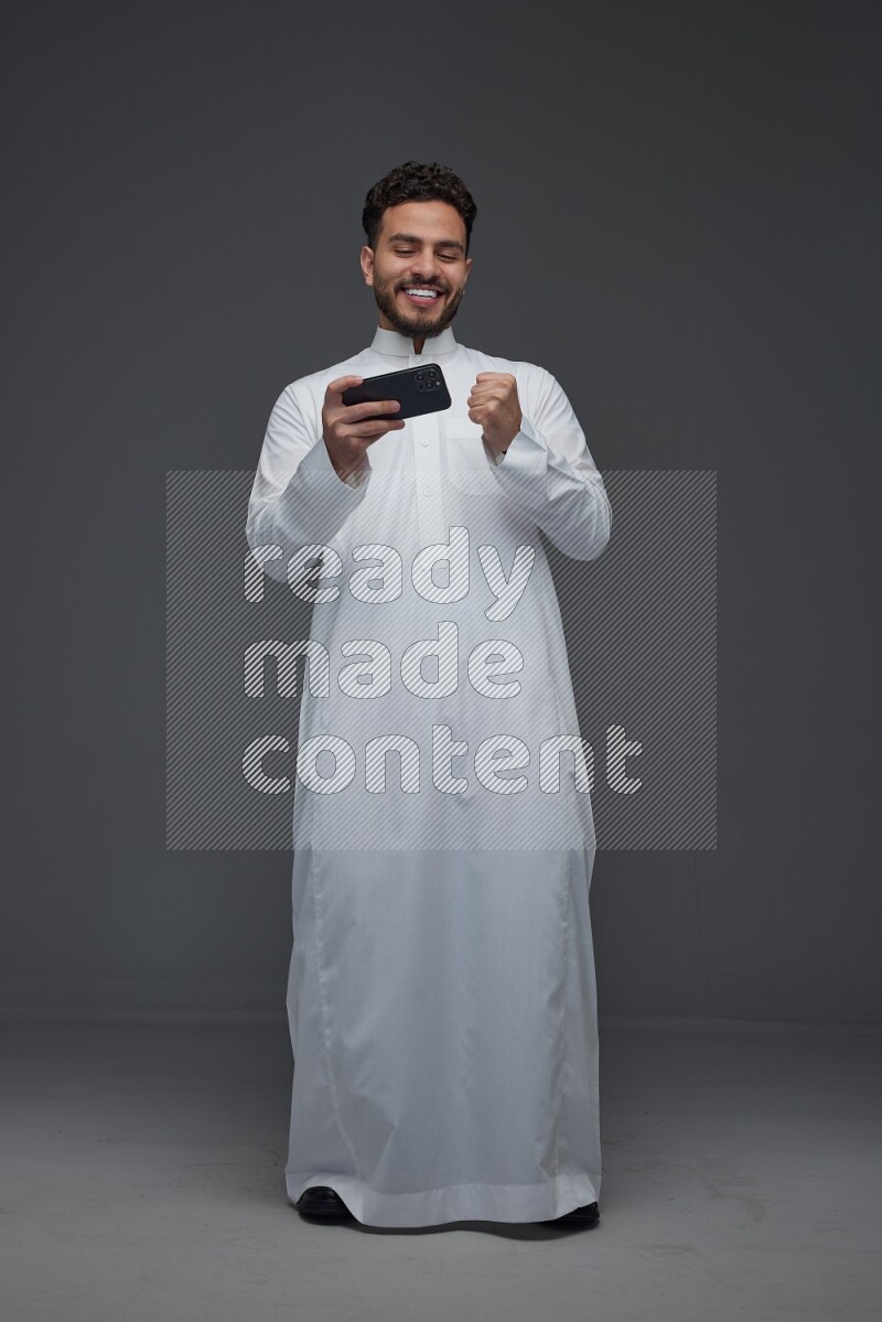 A Saudi man wearing Thobe and playing using his phone horizontally playing video games while standing and making different poses eye level on a gray background