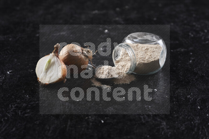 A glass jar full of onion powder flipped with some spilling powder on black background