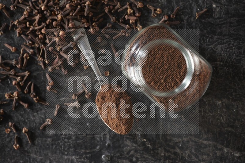 A glass spice jar and a metal spoon full of cloves powder with cloves spread on textured black flooring