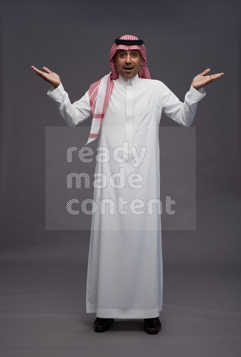Saudi man wearing thob and shomag standing interacting with the camera on gray background
