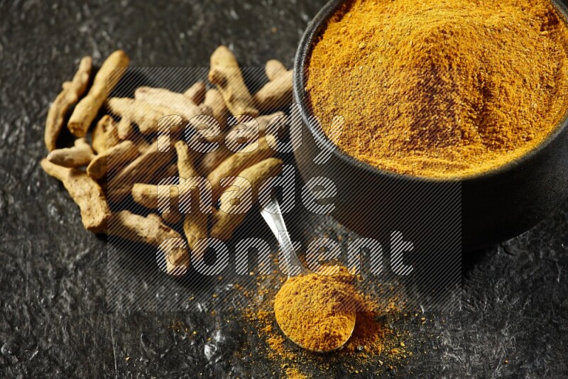 A pottery black bowl and a metal spoon full of turmeric powder with dried turmeric fingers on a textured black flooring