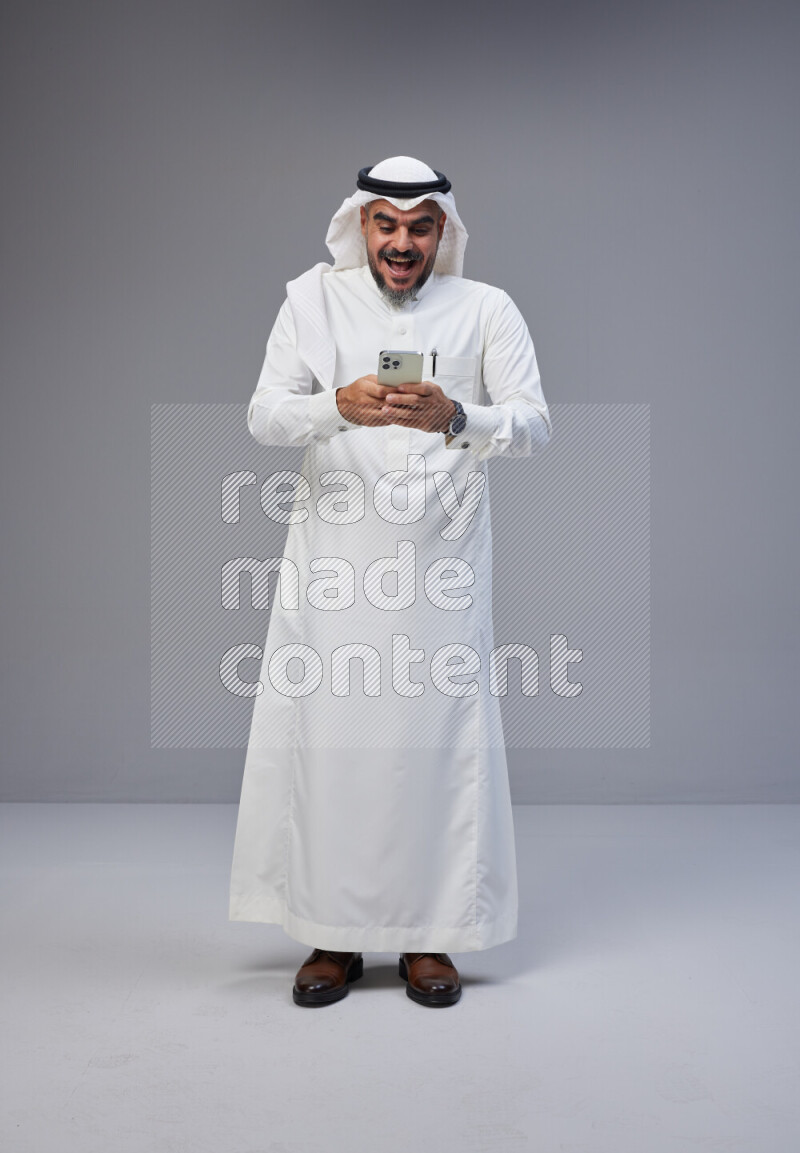 Saudi man Wearing Thob and white Shomag standing texting on phone on Gray background