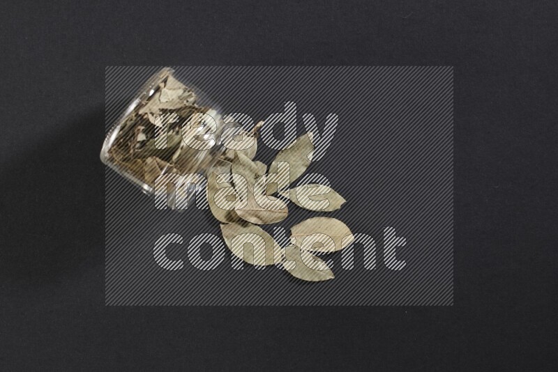 A glass jar full of dried bay leaves and jar is flipped and leaves spread out on black flooring