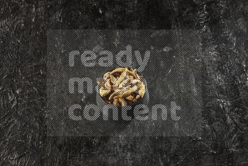 A black pottery bowl full of dried turmeric whole fingers on textured black flooring