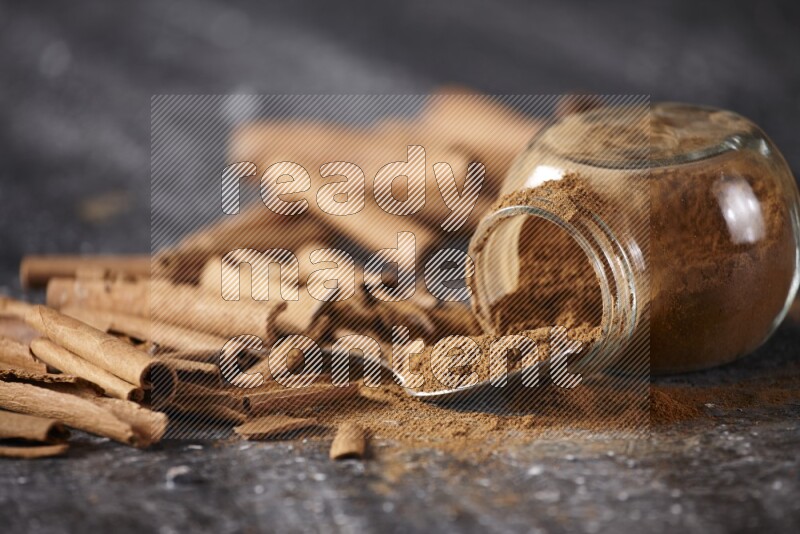 Herbal glass jar full cinnamon powder flipped and a metal spoon full of powder surrounded by cinnamon sticks on textured black background in different angles