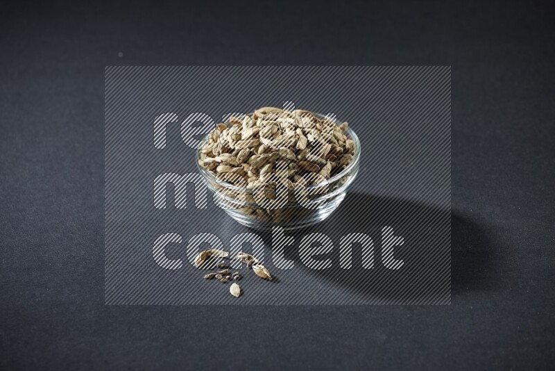 A glass bowl full of cardamom and more seeds spreaded beneath the bowl on black flooring