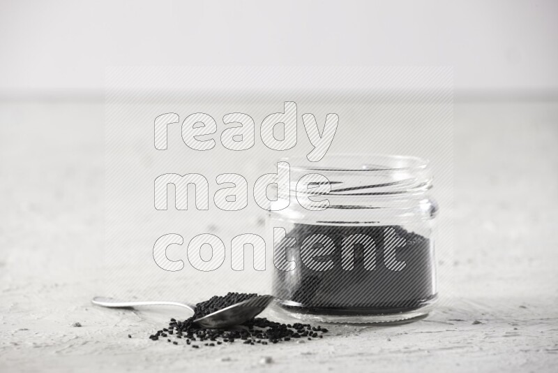 A glass jar and a metal spoon full of black seeds on a textured white flooring