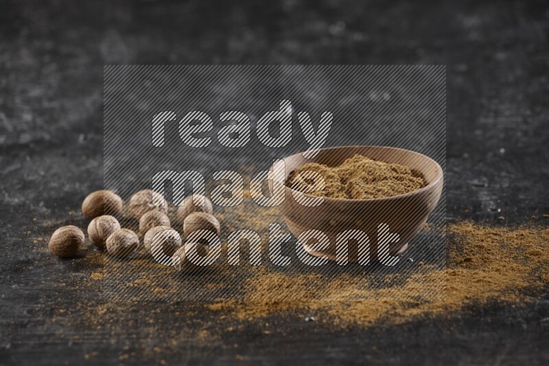 A wooden bowl full of nutmeg powder with whole seeds and sprinkled powder beside it on a textured black flooring