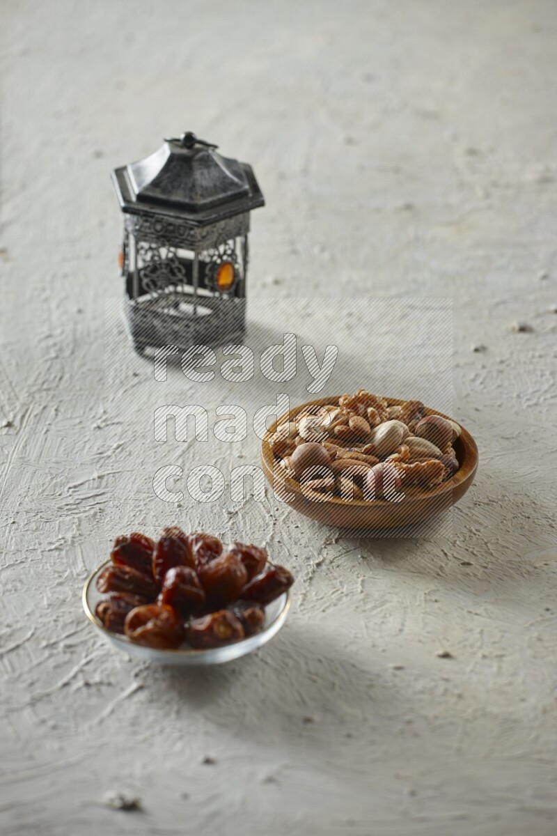 A silver lantern with different drinks, dates, nuts, prayer beads and quran on textured white background