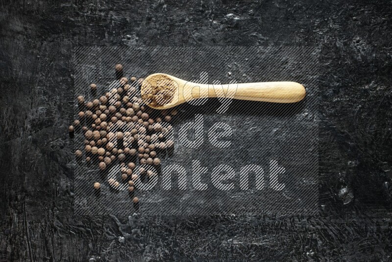 A wooden spoon full of allspice powder and whole balls spreaded on a textured black flooring