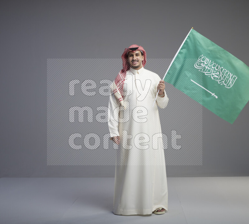 A Saudi man standing wearing thob and red shomag raising big Saudi flag on gray background