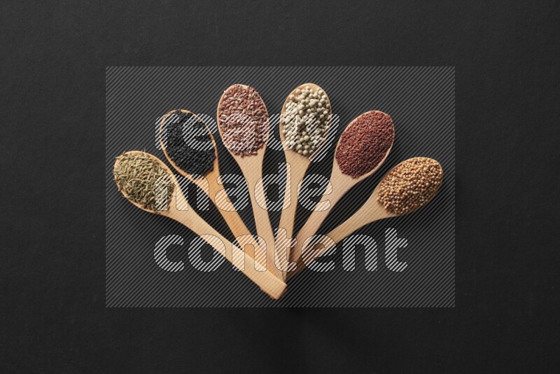 wooden spoons filled with white peppers, mustard seeds, black seeds, garden cress, cumin and flax on black flooring