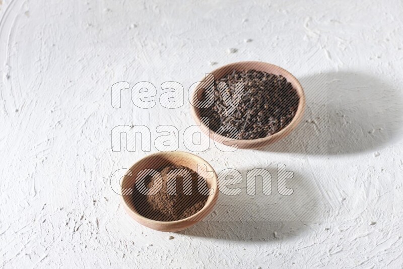 2 wooden bowls full of cloves powder and whole cloves on a textured white flooring