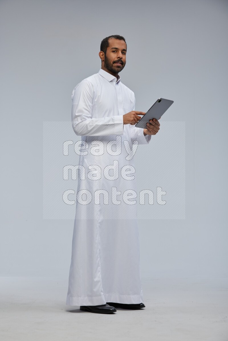 Saudi man wearing Thob standing working on tablet on Gray background