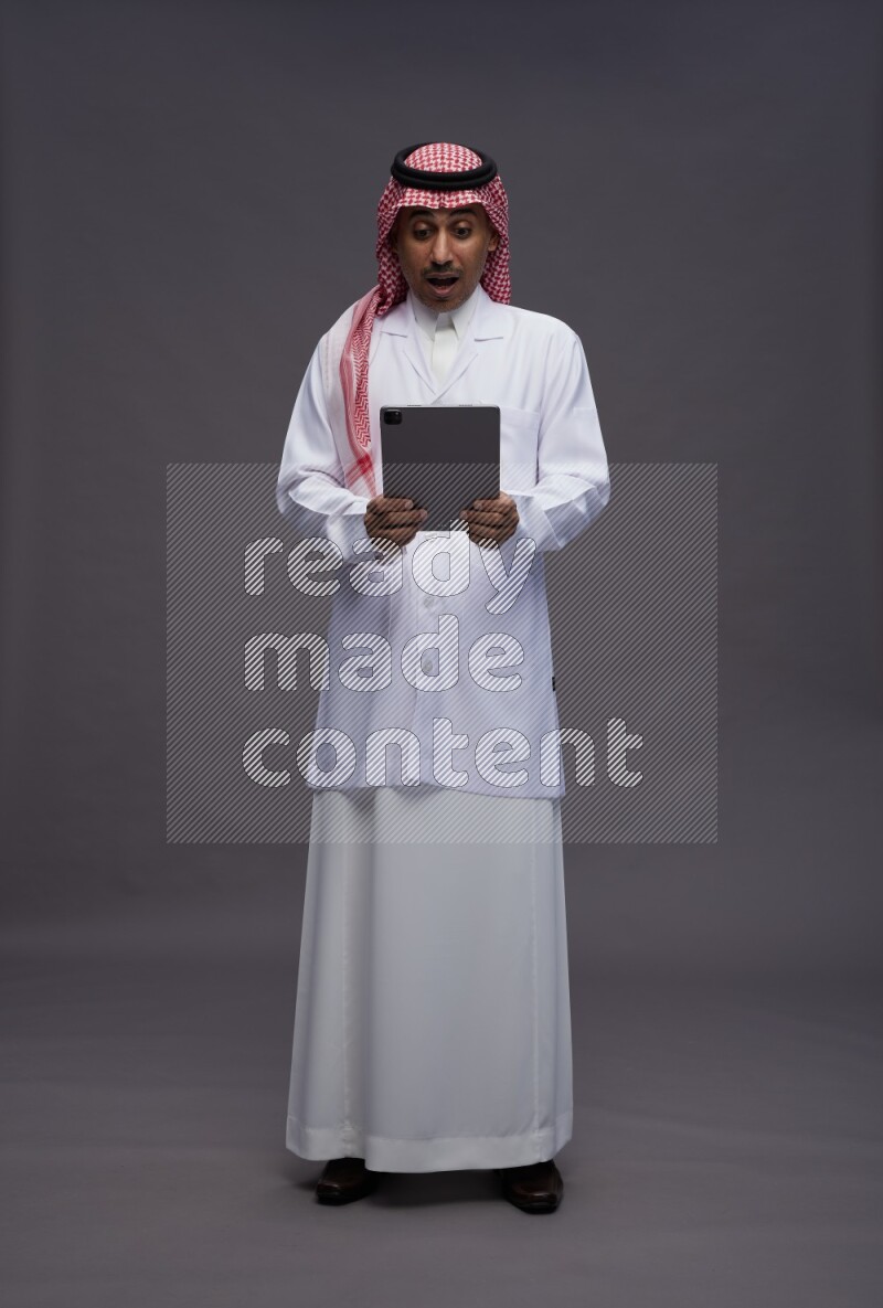 Saudi man wearing thob with lab coat and shomag with pocket employee badge standing working on tablet on gray background