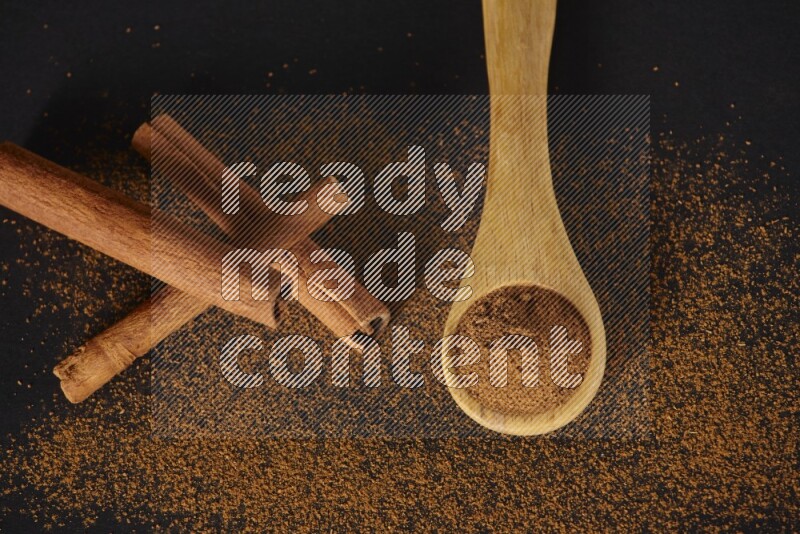 Cinnamon powder in a wooden spoon and cinnamon sticks beside it on black background