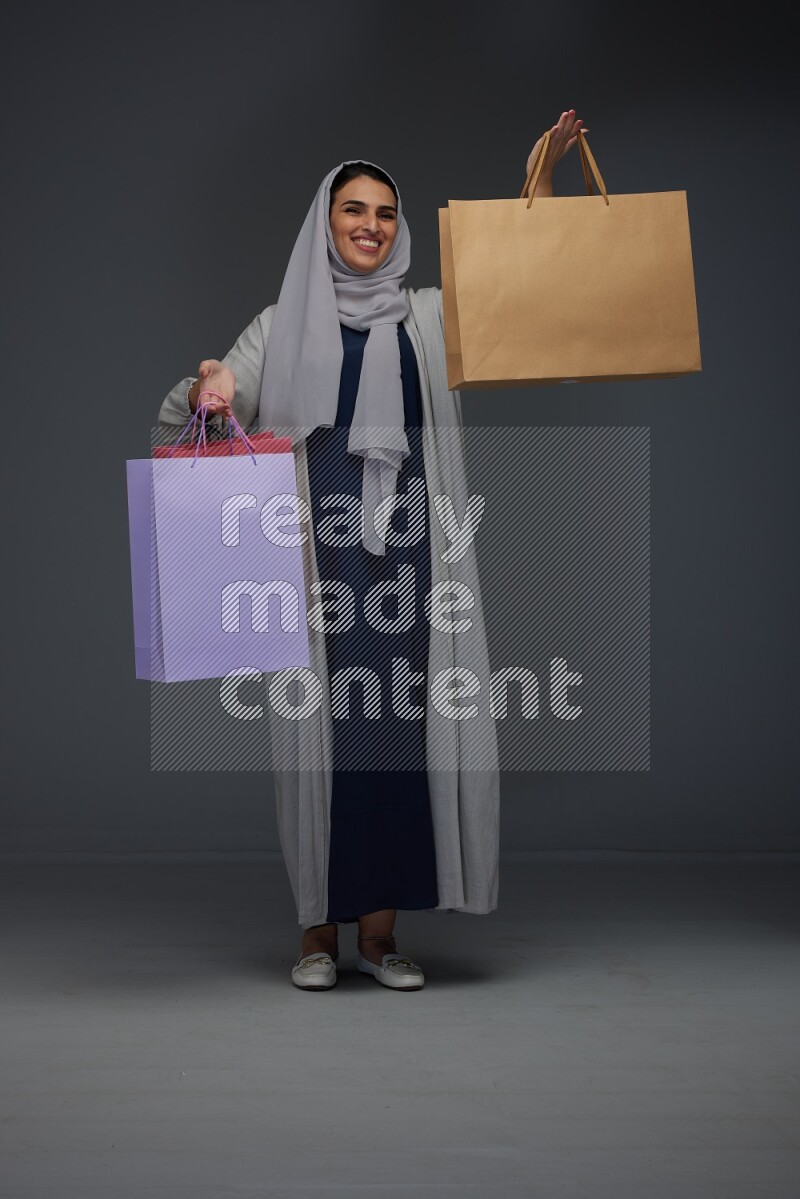 A Saudi woman wearing a light gray Abaya and head scarf standing and holding shopping bags on a grey background