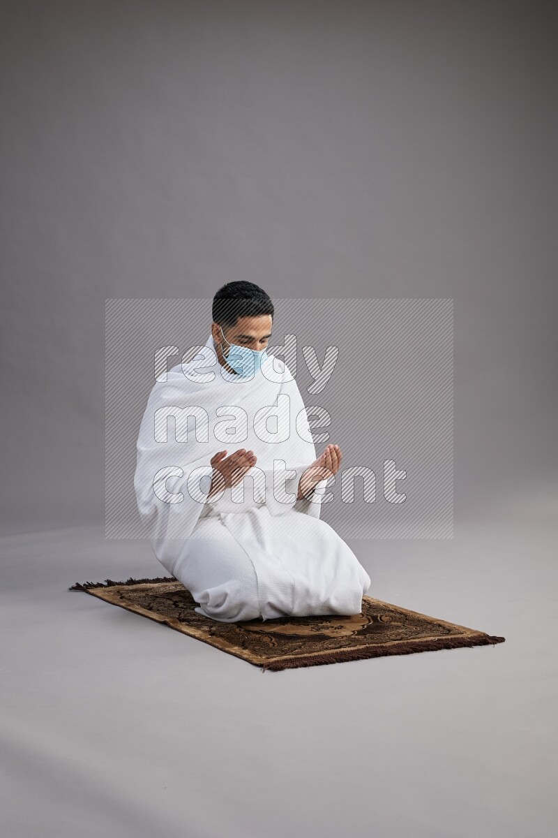 A man wearing Ehram with face mask sitting on floor performing dua'a on gray background