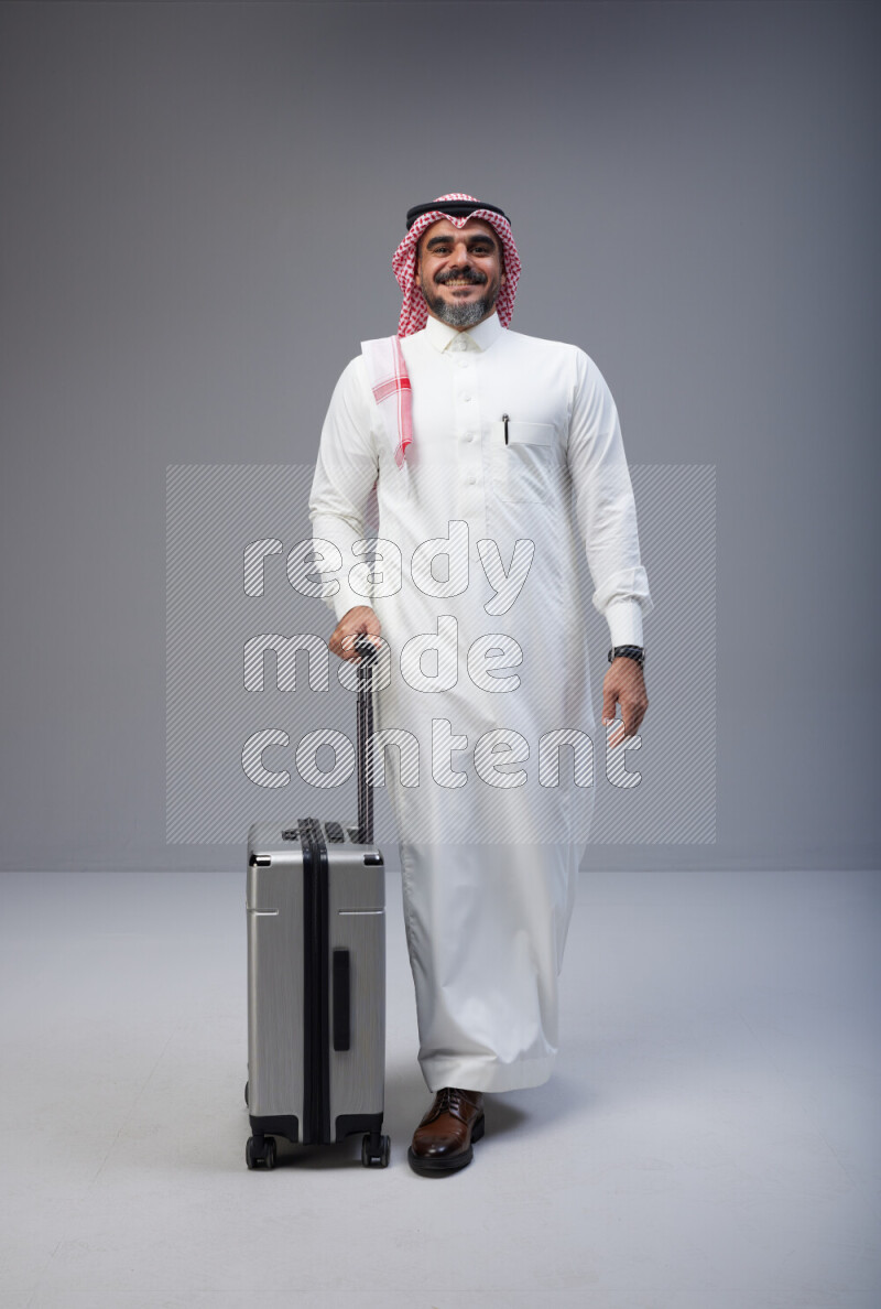 Saudi man wearing Thob and red Shomag standing holding Travel bag on Gray background