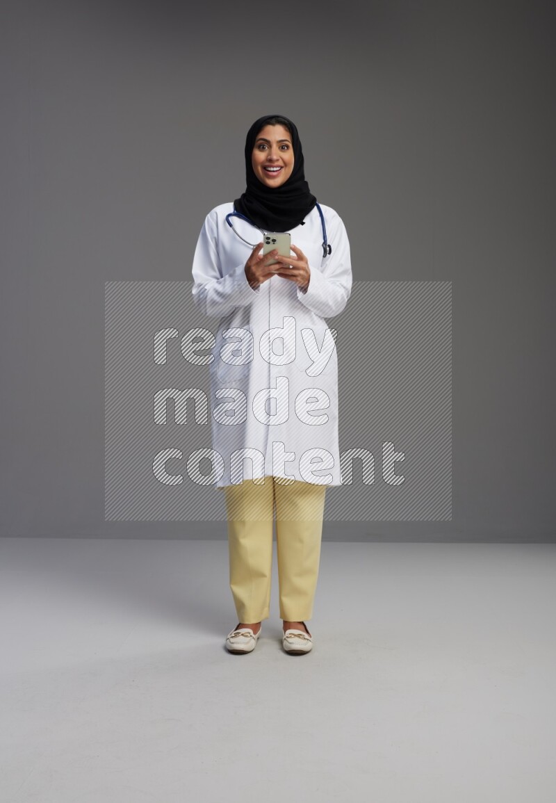Saudi woman wearing lab coat with stethoscope standing texting on phone on Gray background