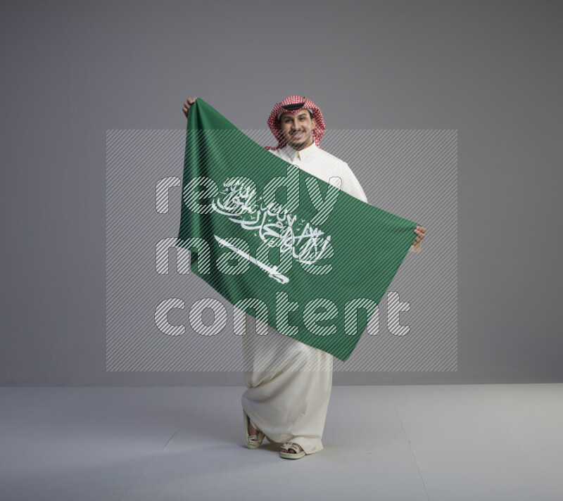 A saudi man standing wearing thob and red shomag holding big saudi flag on gray background