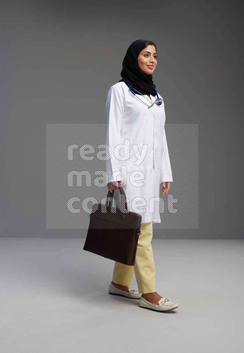 Saudi woman wearing lab coat with stethoscope standing holding bag on Gray background