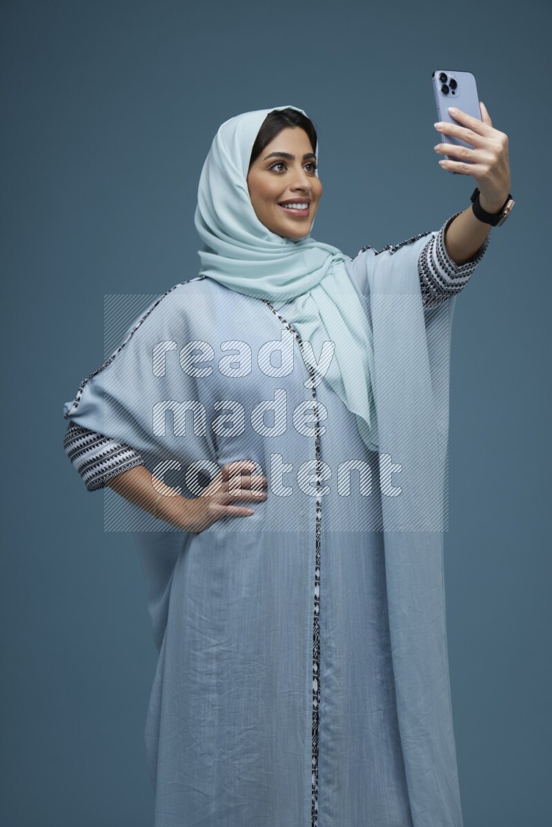 A Saudi woman Taking a Selfie on a blue background wearing a blue Abaya with hijab