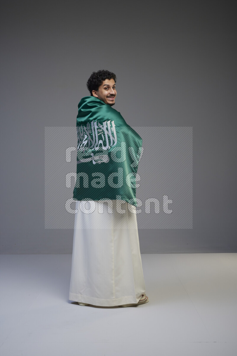 A Saudi man standing wearing thob wrapping big Saudi flag on gray background