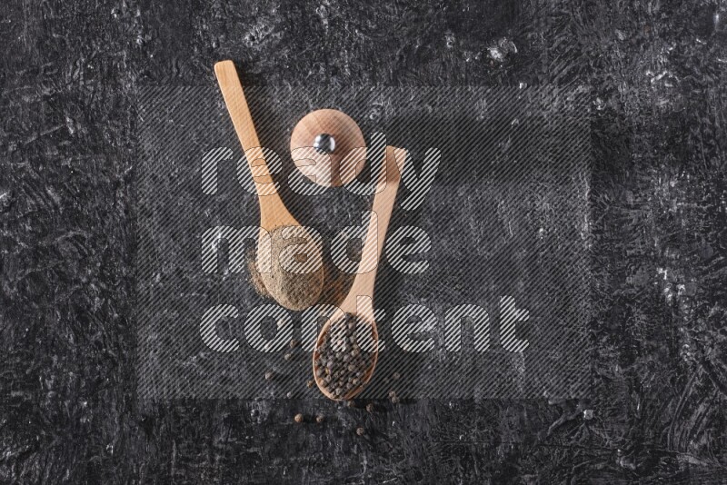 Black pepper powder and black pepper beads in wooden spoons and wooden grinder on a textured black flooring