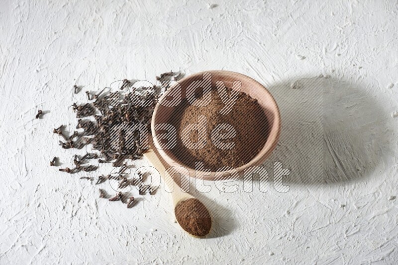 A wooden bowl and wooden spoon full of cloves powder with cloves spread on textured white flooring