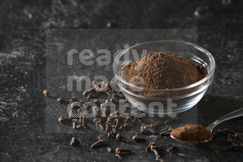 A glass bowl and a metal spoon full of cloves powder with gloves grains beside them on a textured black flooring