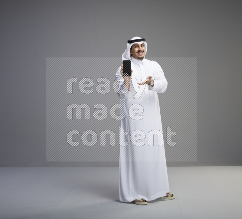 A Saudi man standing wearing thob and white shomag showing phone to camera on gray background