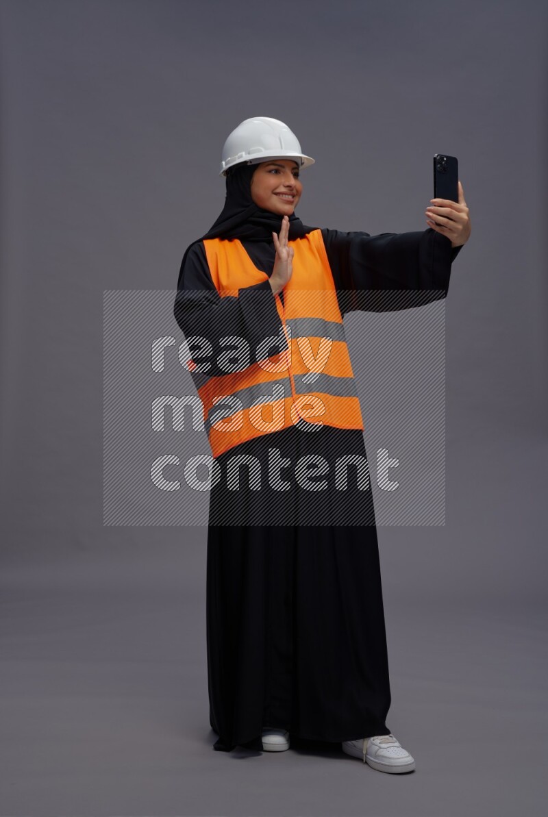 Saudi woman wearing Abaya with engineer vest standing taking selfie on gray background