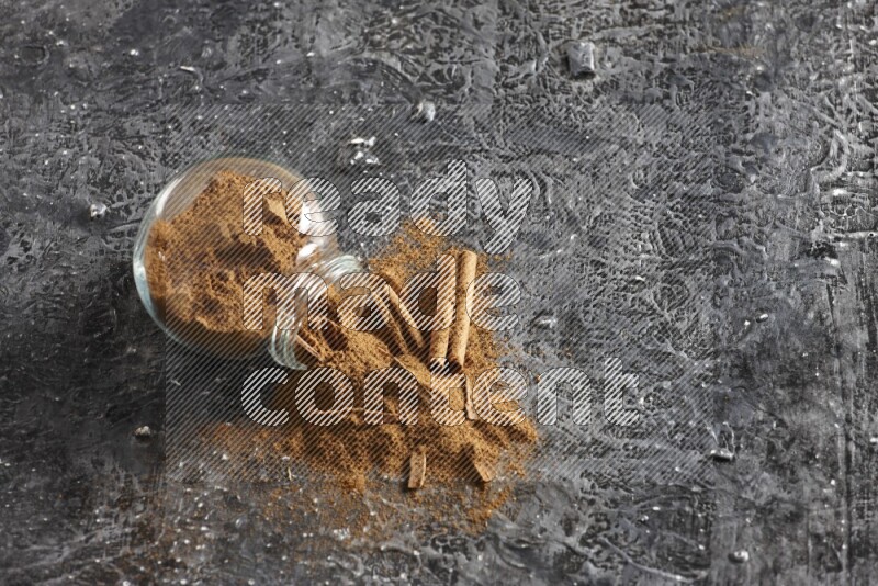 Flipped glass herbs jar full of cinnamon powder with cracked cinnamon sticks on a textured black background