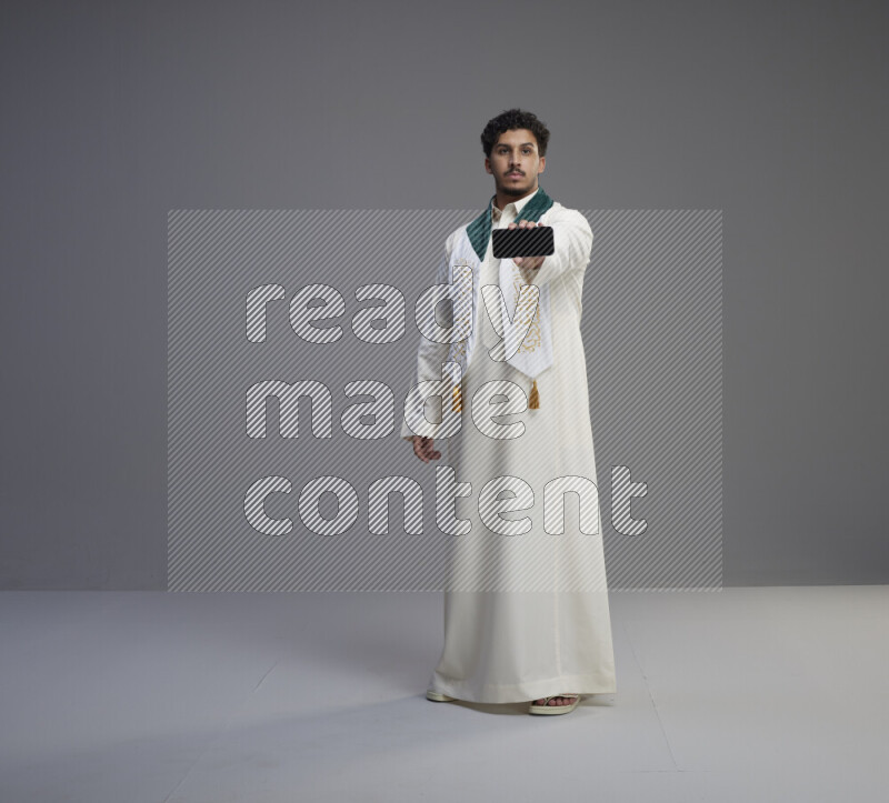 A Saudi man standing wearing thob and Saudi flag scarf showing phone to camera on gray background