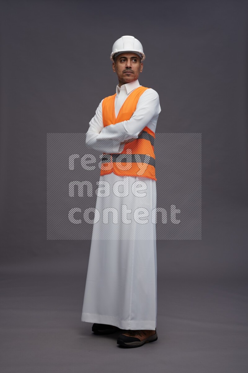 Saudi man wearing thob with engineer vest standing with crossed arms on gray background