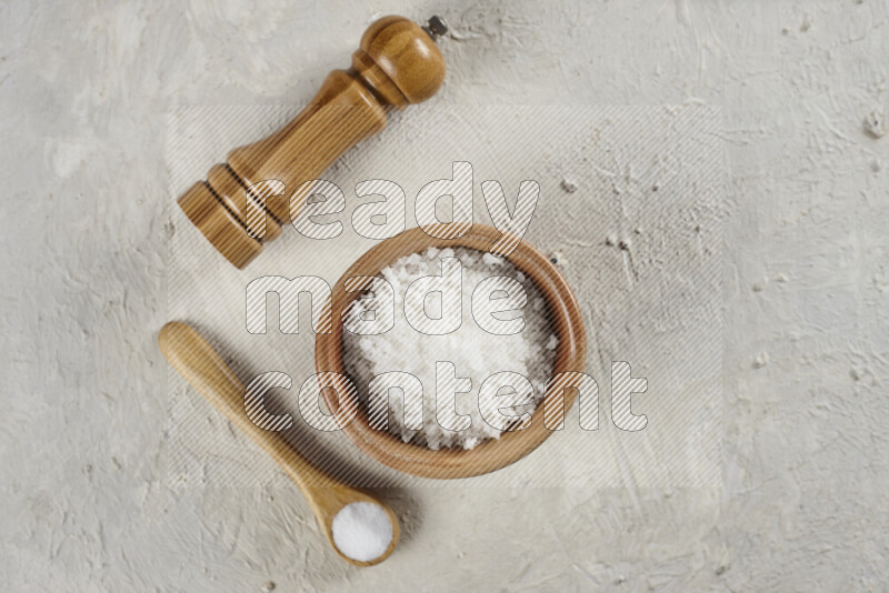 A wooden bowl and spoon filled with white sea salt and wooden grinder beside them on white background
