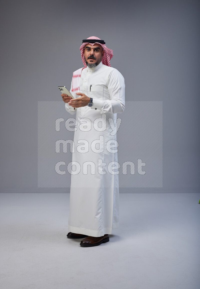 Saudi man Wearing Thob and red Shomag standing texting on phone on Gray background