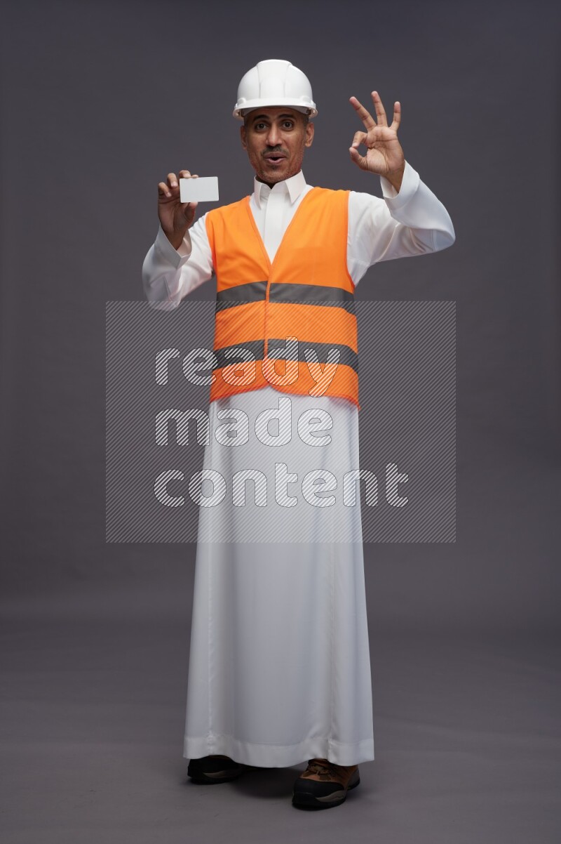 Saudi man wearing thob with engineer vest standing holding ATM card on gray background