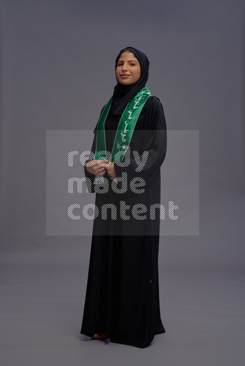 Saudi woman wearing Abaya standing holding Saudi flag on gray background
