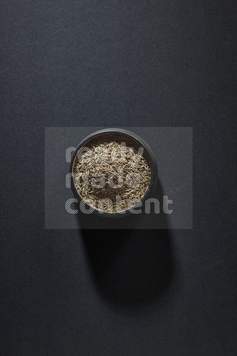 A black pottery bowl full of cumin seeds on a black flooring
