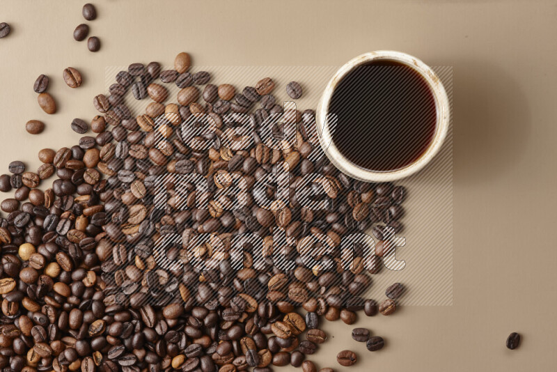 A beige pottery cup of coffee surrounded by roasted coffee beans on beige background