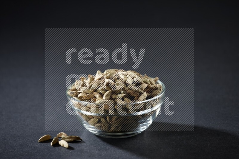 A glass bowl full of cardamom and more seeds spreaded beneath the bowl on black flooring