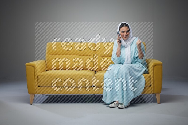 A Saudi woman wearing a light blue Abaya and a white head scarf sitting on a yellow sofa and talking in the phone while making different poses eye level on a grey background