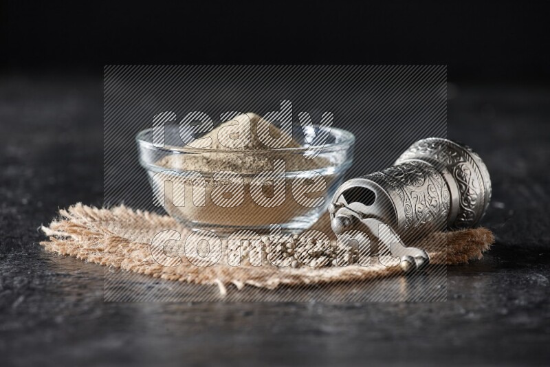 A glass bowl full of white pepper powder with white pepper beads on a burlap piece of fabric and a metal grinder on textured black flooring